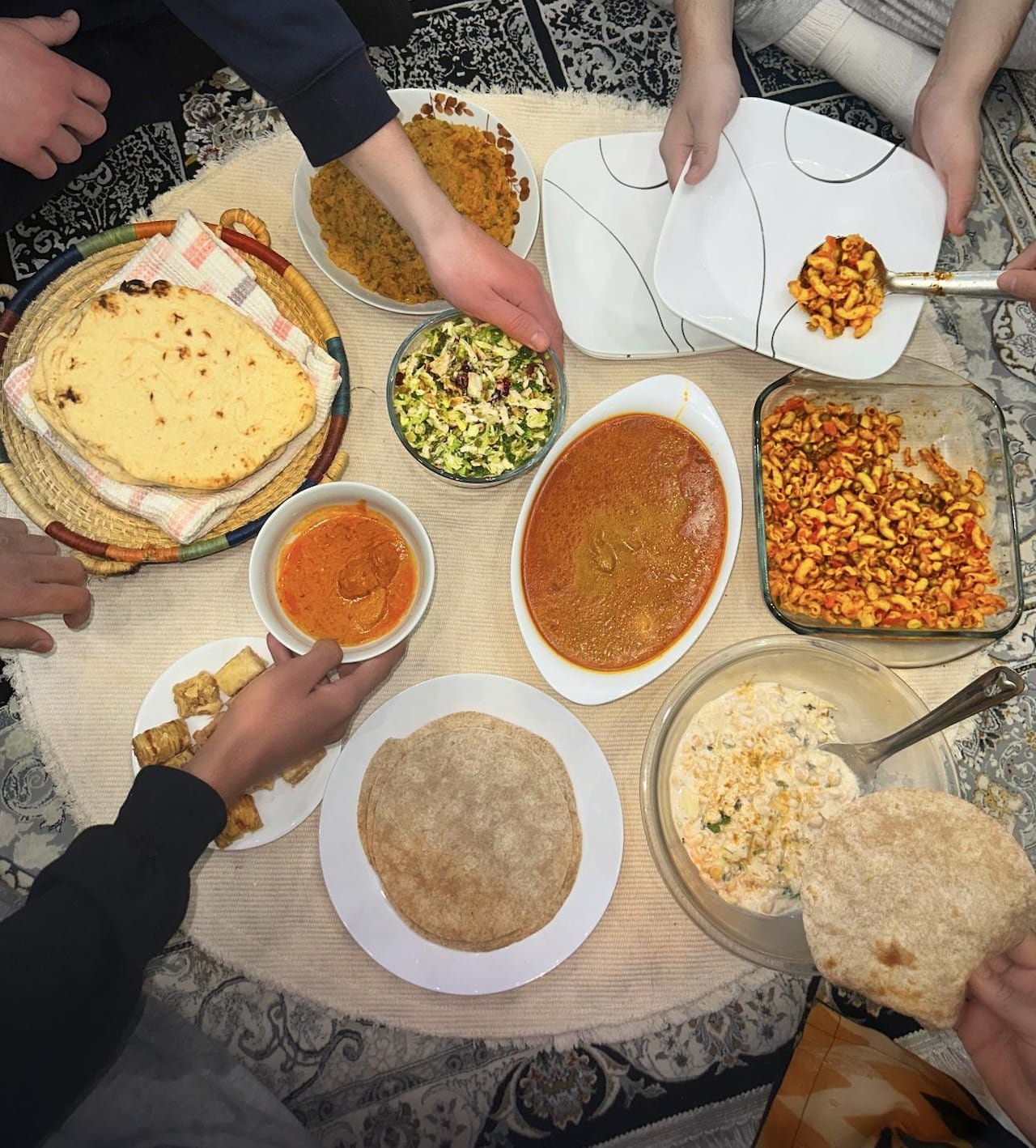 default-235 A bird’s eye view of several hands reaching for food, such as butter chicken, macaroni salad, roti and baklava.