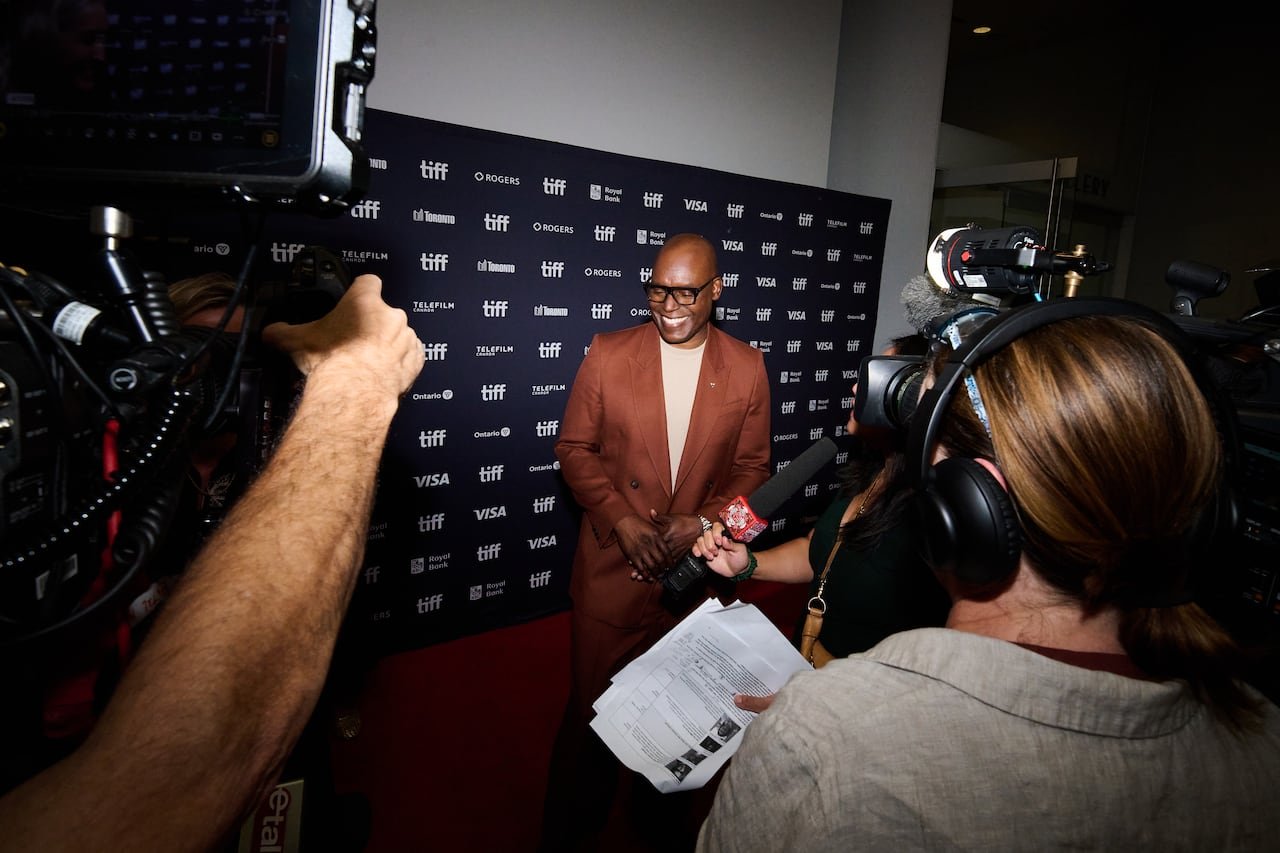 default-113 A man wearing a brown suit and glasses stands on the TIFF red carpet with cameras around him, with a black screen background behind him bearing the words "TIFF."